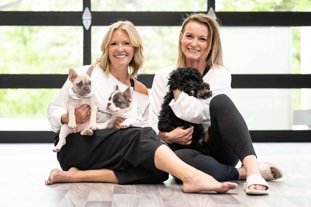 Two smiling Downward Dogs Puppy Yoga instructors holding puppies in a sunlit studio